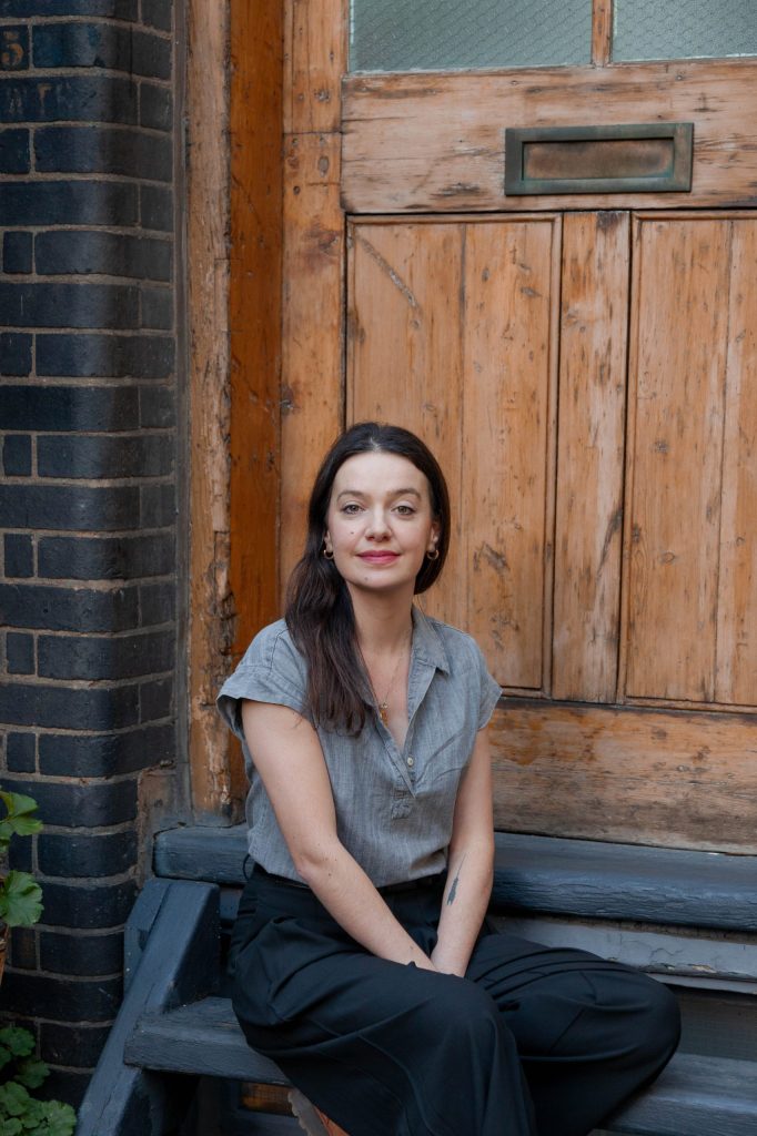 Photo of Rebecca King sat on a wooden step outside a door.